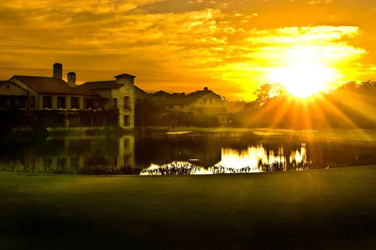 The Bear’s Club clubhouse at sunset, its stone facade reflected in the lake beyond the 18th green