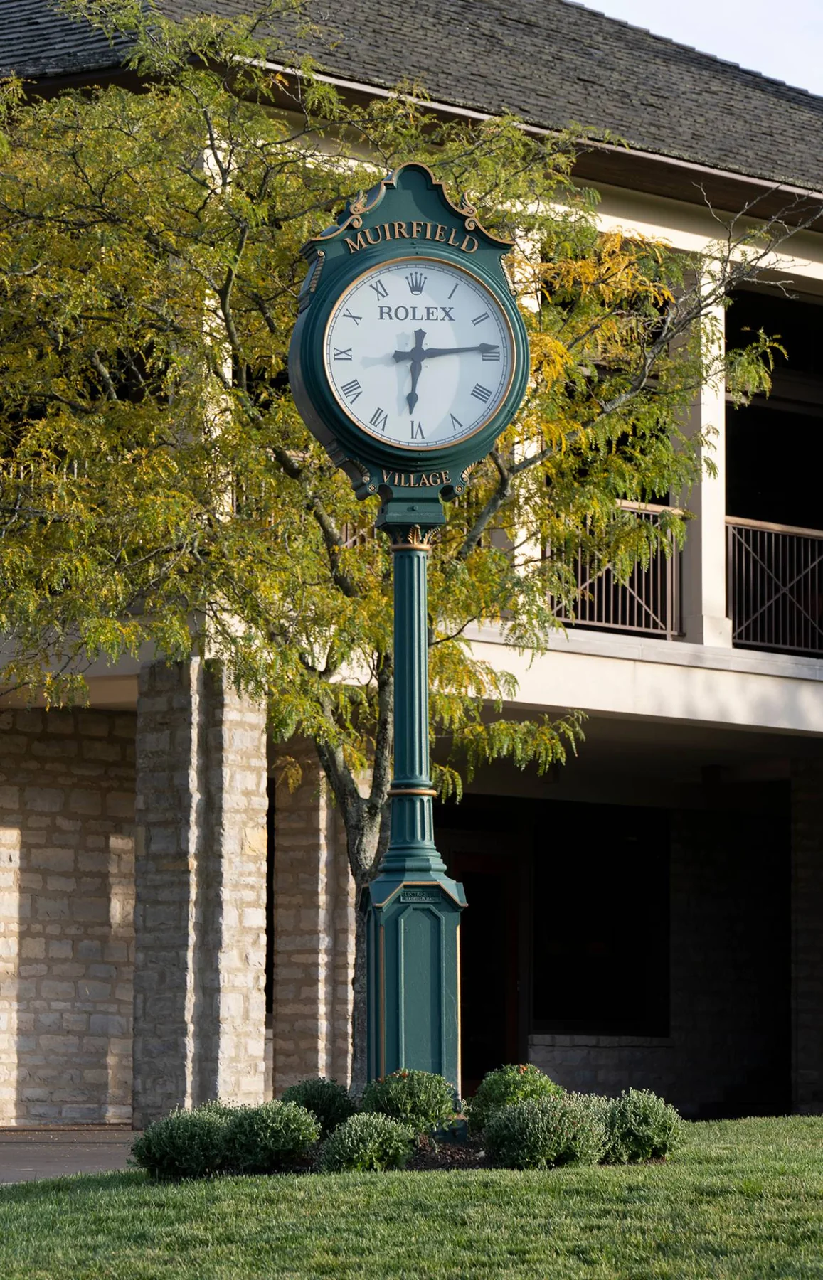 A green and gold Rolex post clock reading Muirfield Village in front of the clubhouse