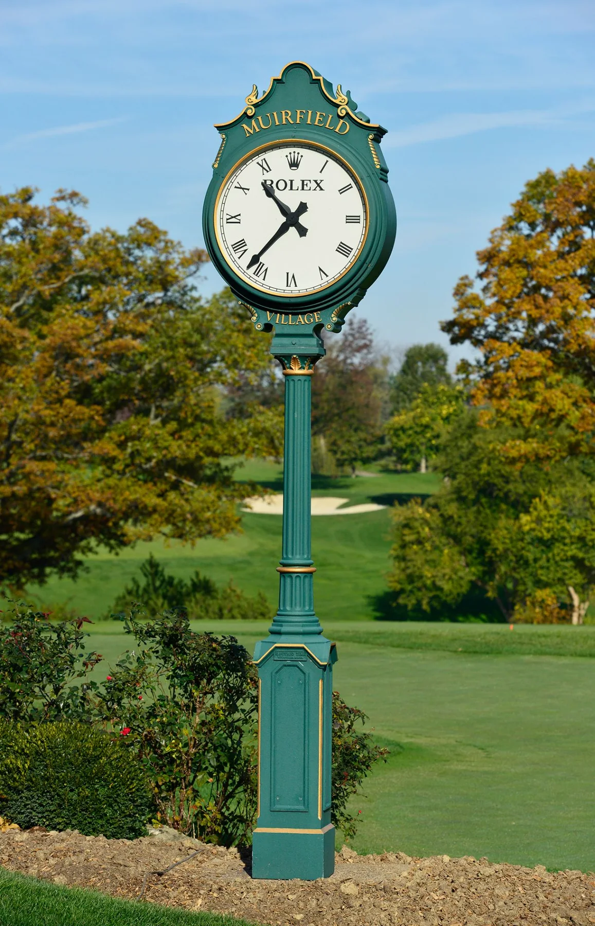 A green and gold Rolex post clock reading Muirfield Village, the course and a bunker behind it